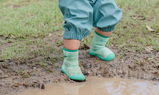 Toddler in green wide toe wee wellies and waterproof rain pants, cautiously stepping into a puddle