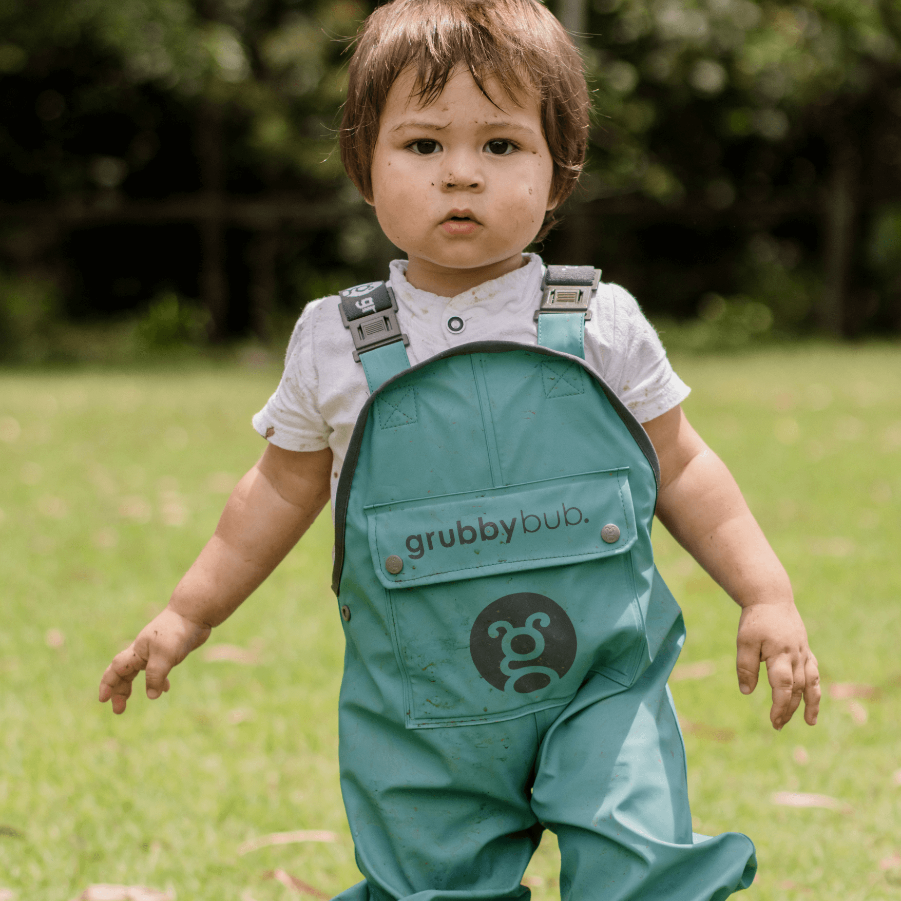 A child wearing green waterproof overalls with adjustable straps standing on grass.