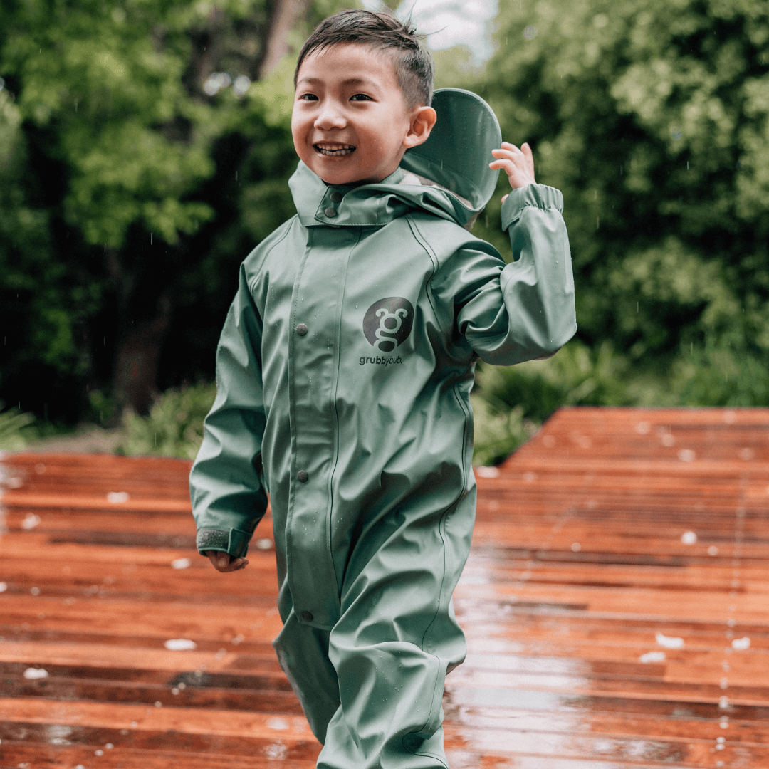 Asian boy in grubbybub sage green puddle suit on a wet deck 