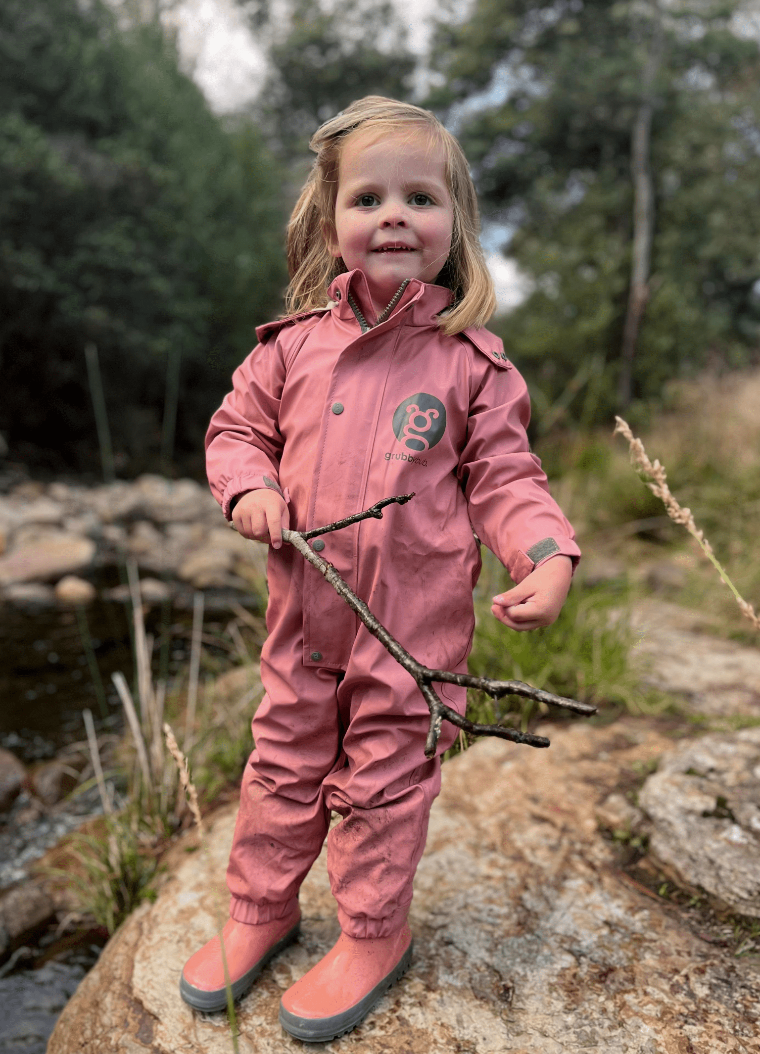 toddler girl in dusty rose pink waterproof rain suit and adjustable gumboots, standing by a creek holding a stick