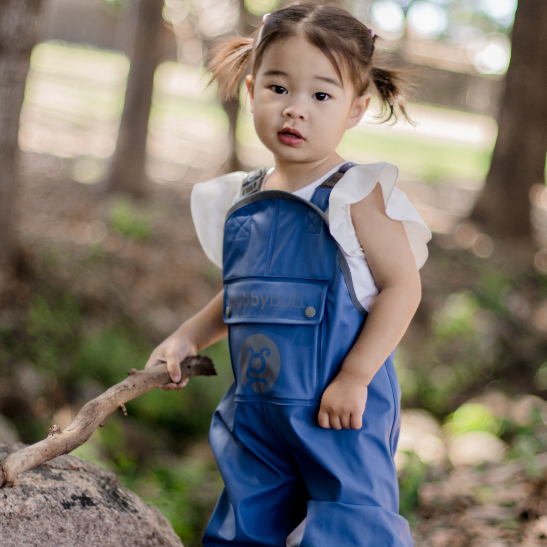 Child in blue sustainle waterproof overalls holding a stick in a forest setting