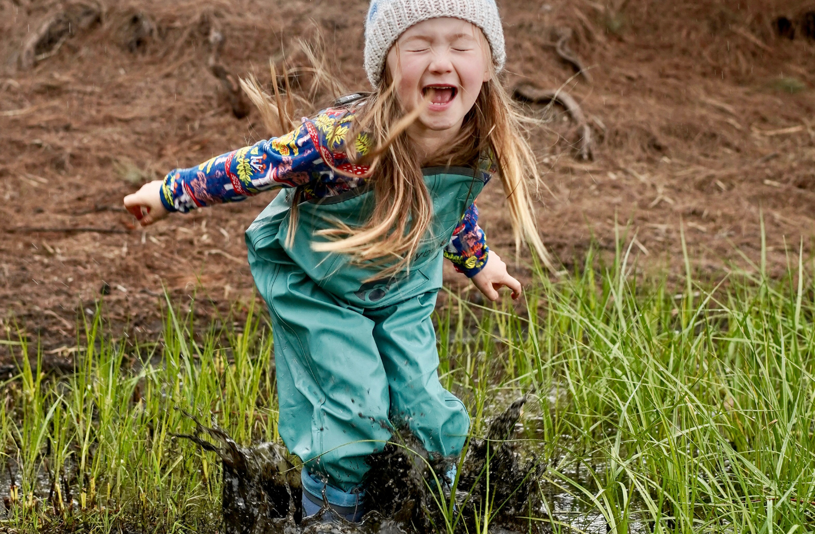 A long haired happy toddler girl wearing deep sea green waterproof grubberall wader pants and gumboots makes a huge muddy splash by jumping