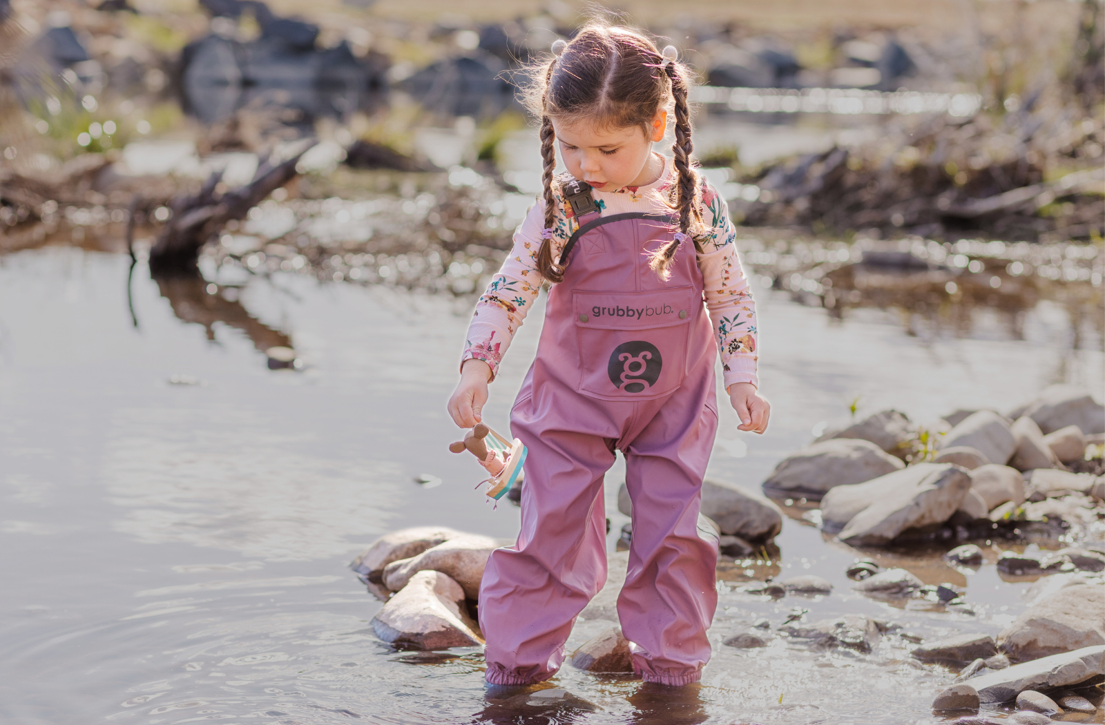 A brown haired toddler girl playing ankle deep in a lake, wearing her waterproof grubberall rain pants in plum purple colour 