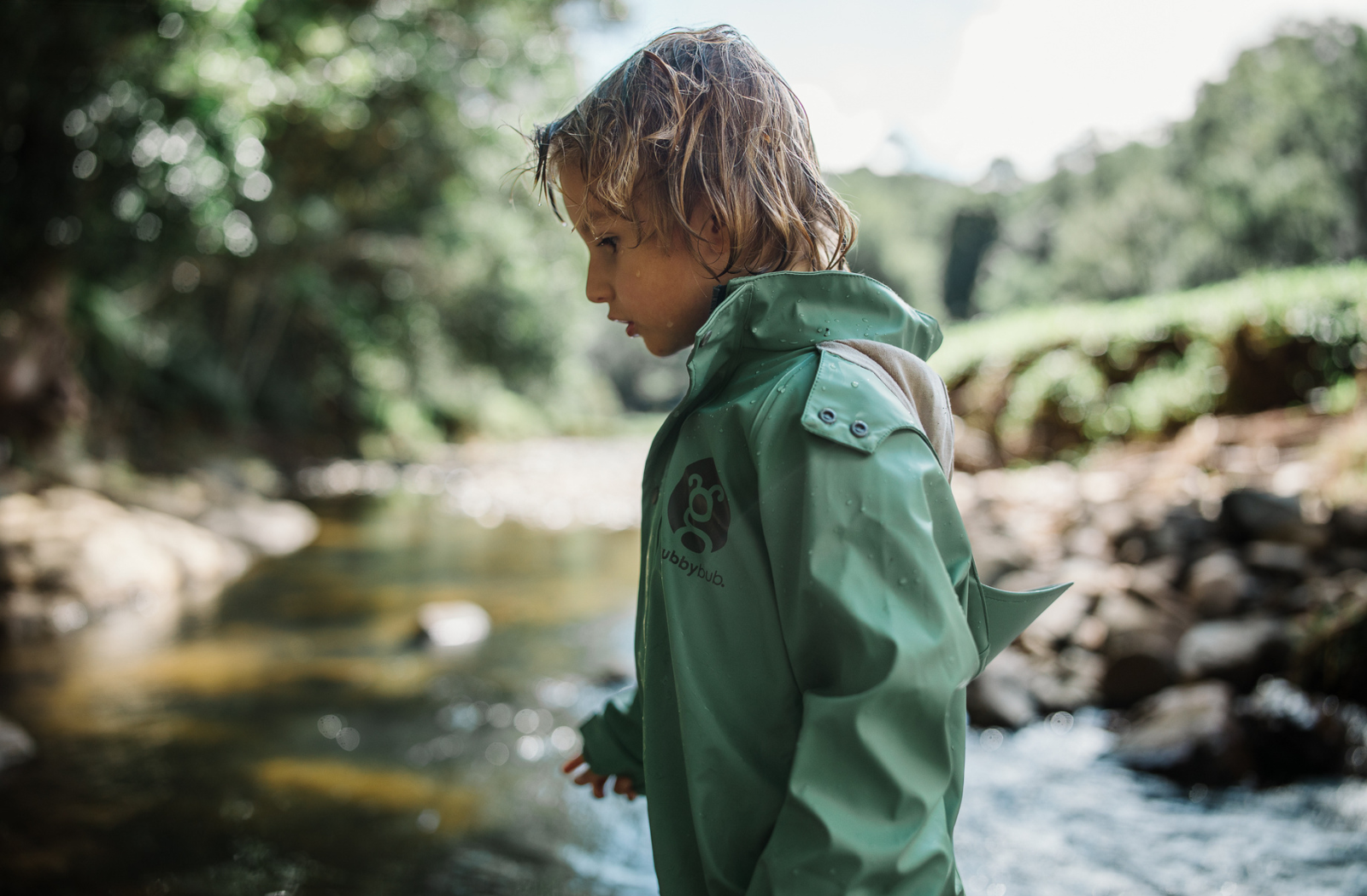 A toddler boy wearing a sage green waterproof puddle suit exploring a creek