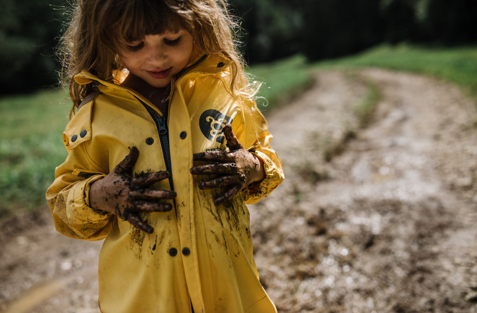 Brown haired toddler girl wiping her muddy hands on her waterproof sunshine yellow puddle suit