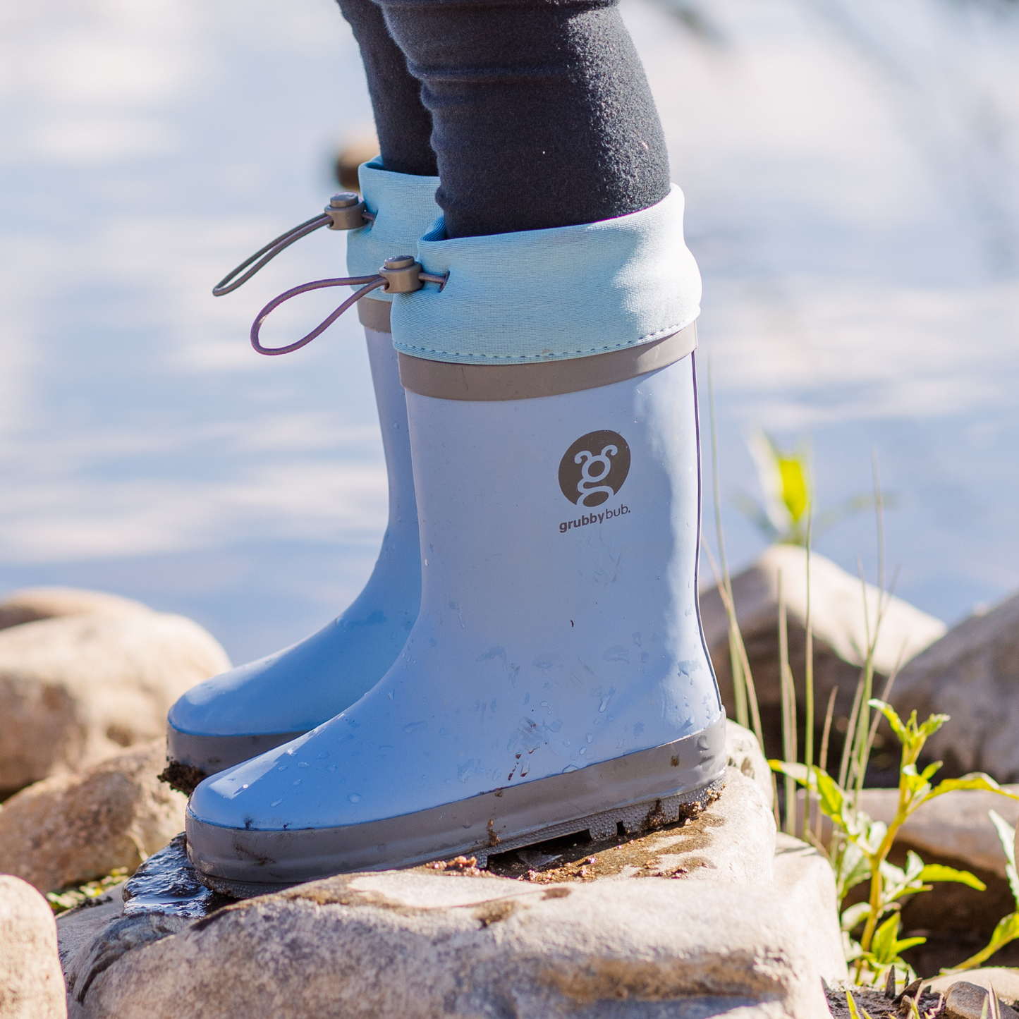 Blue rubber boots with an ajustable calf opening on a rocky surface near water.