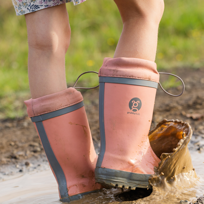 Person wearing adjustable topped pink rubber boots with a visible brand logo, stepping into a puddle outdoors.