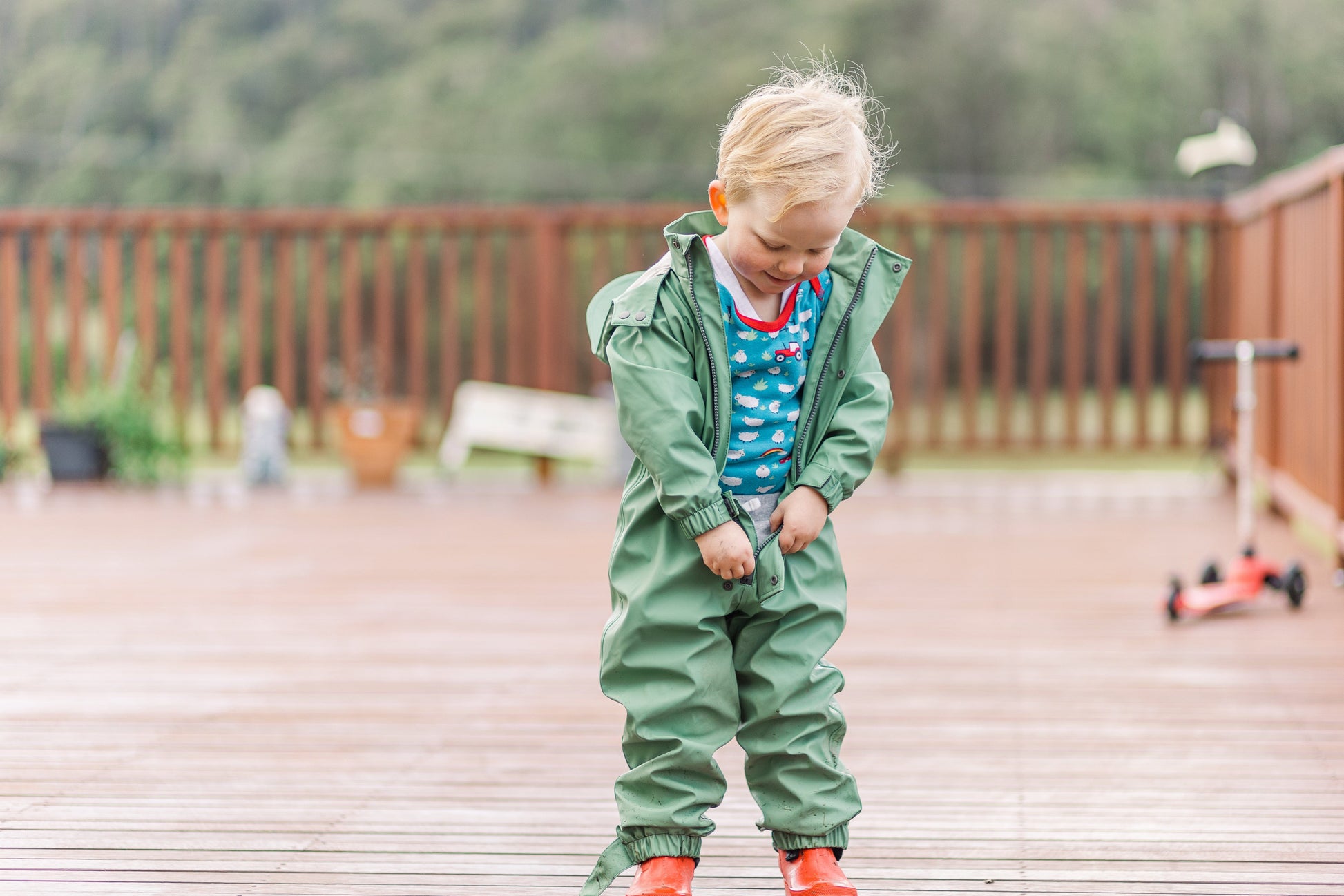 A happy toddler boy in a sage green puddle suit undoing his suit to display his dry clothes underneath