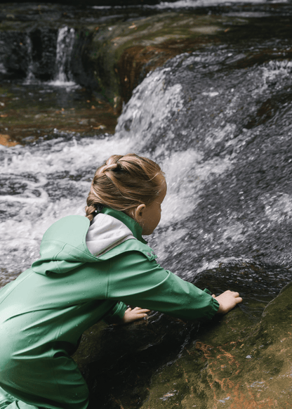 A girl in a sage green puddle suit with her hands in a waterfall
