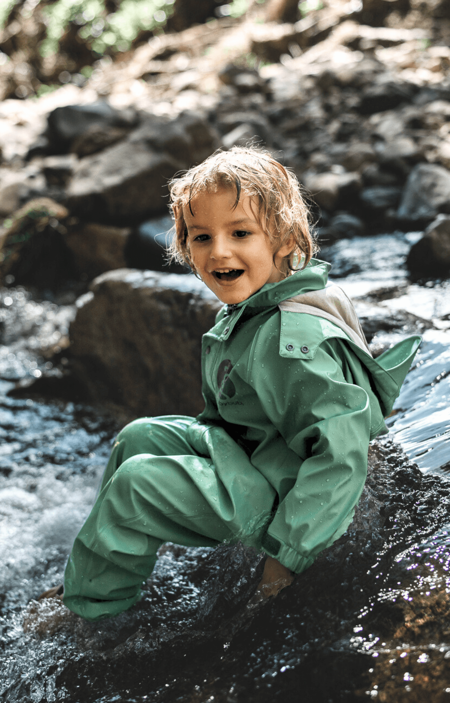 Child wearing green waterproof suit sitting in creek during outdoor water play