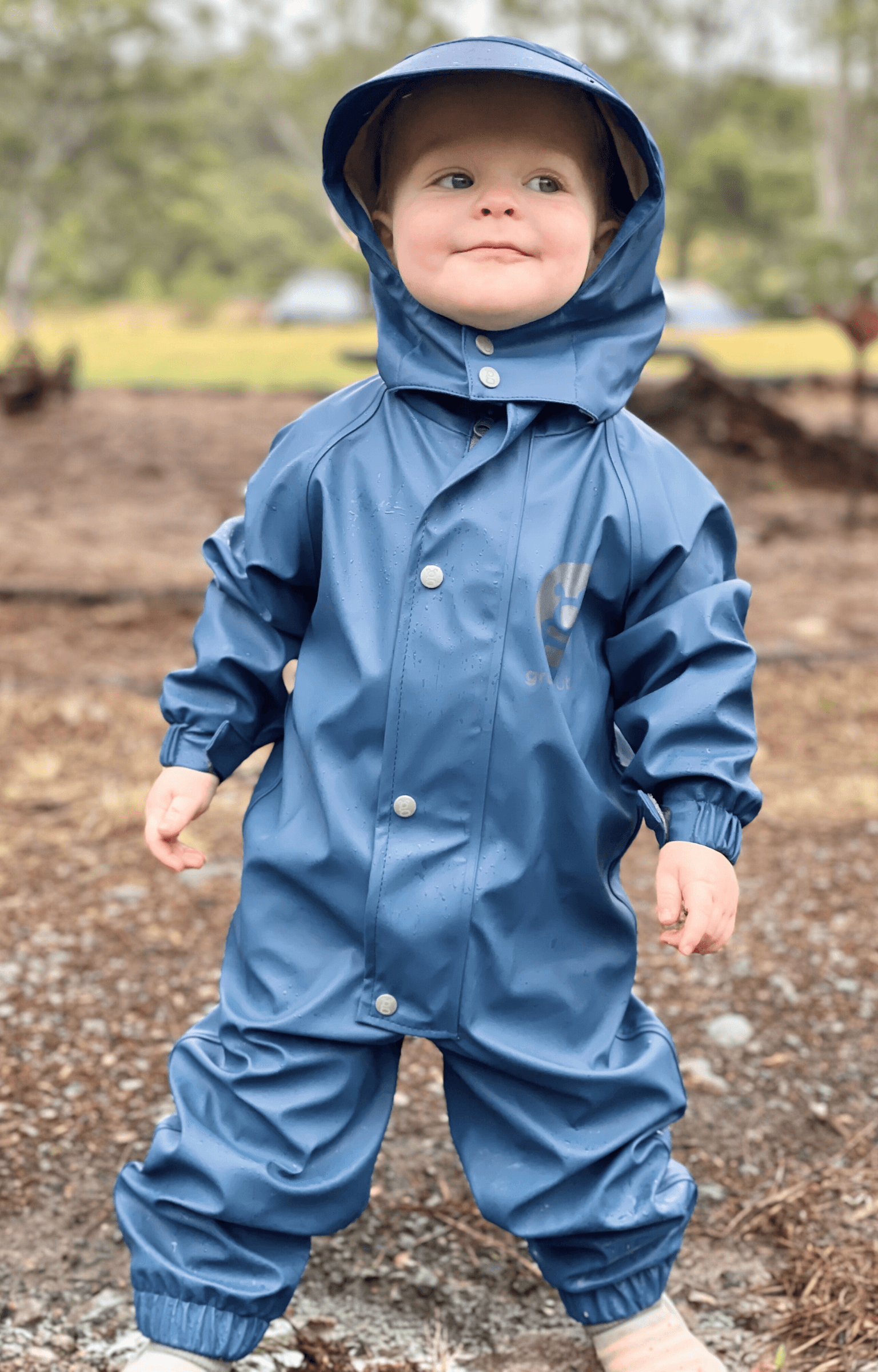 Toddler wearing blue waterproof rain suit standing outdoors, durable kids all weather playwear for forest school and camping